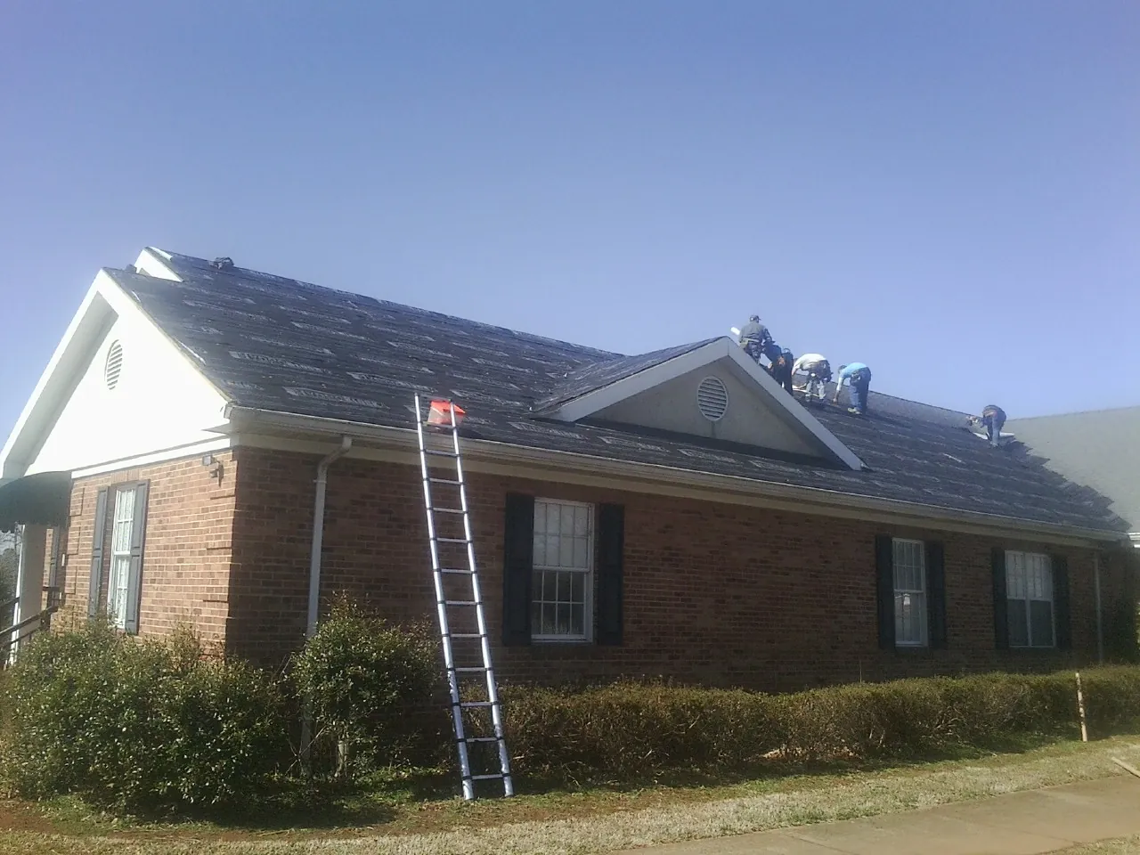 Crew tearing shingles off a brick ranch roof with a ladder set — Metal Roof Repair work by our crew in Grimes