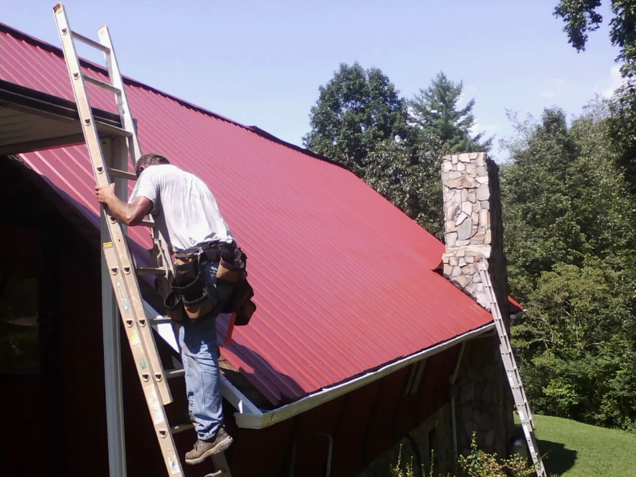 Roofer on a ladder beside a red metal roof and stone chimney — Soffit & Fascia Repair by Roofing Brothers in Grimes